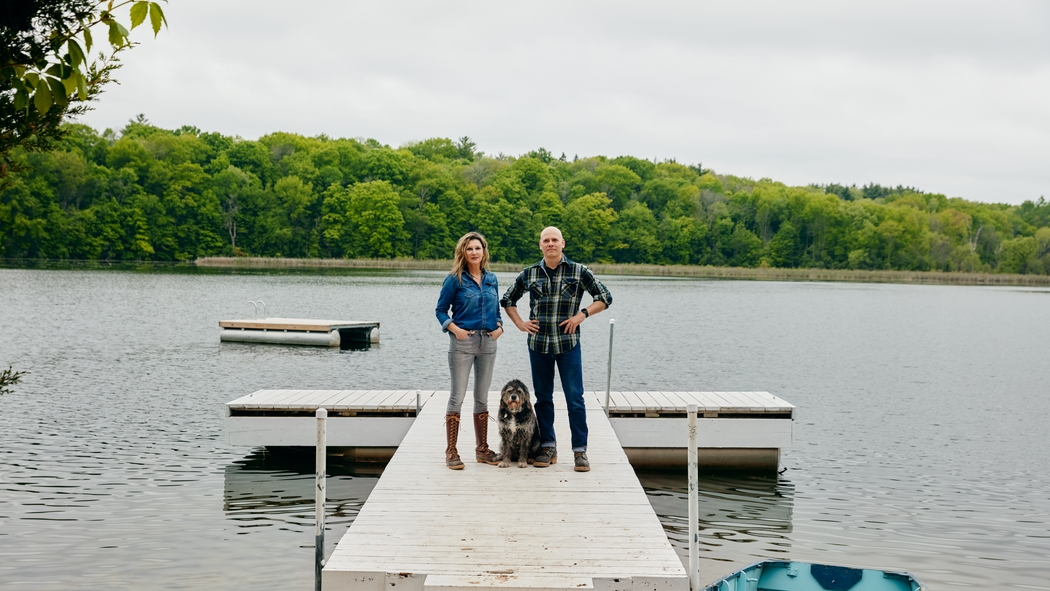 A couple stand with a dog between them on a dock surrounded by water with a rowboat.