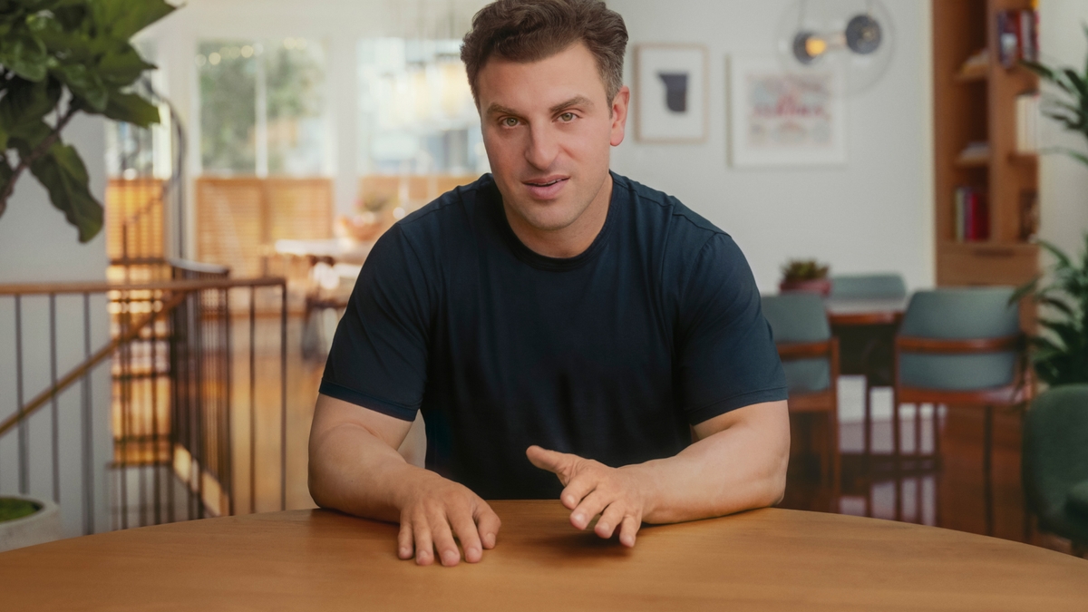Tummaan t-paitaan pukeutunut Airbnb:n toimitusjohtaja Brian Chesky nojaa käsillään puista pöytää vasten ja katsoo kameraan.