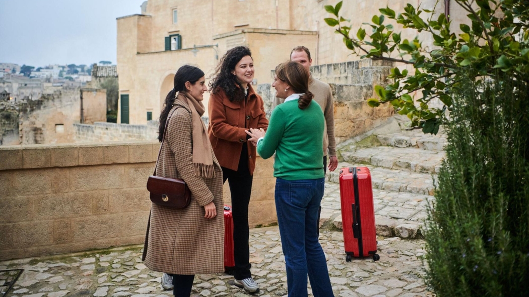 A Host shakes hands with one of three guests standing beside suitcases on a cobblestone walkway tinged green with moss.