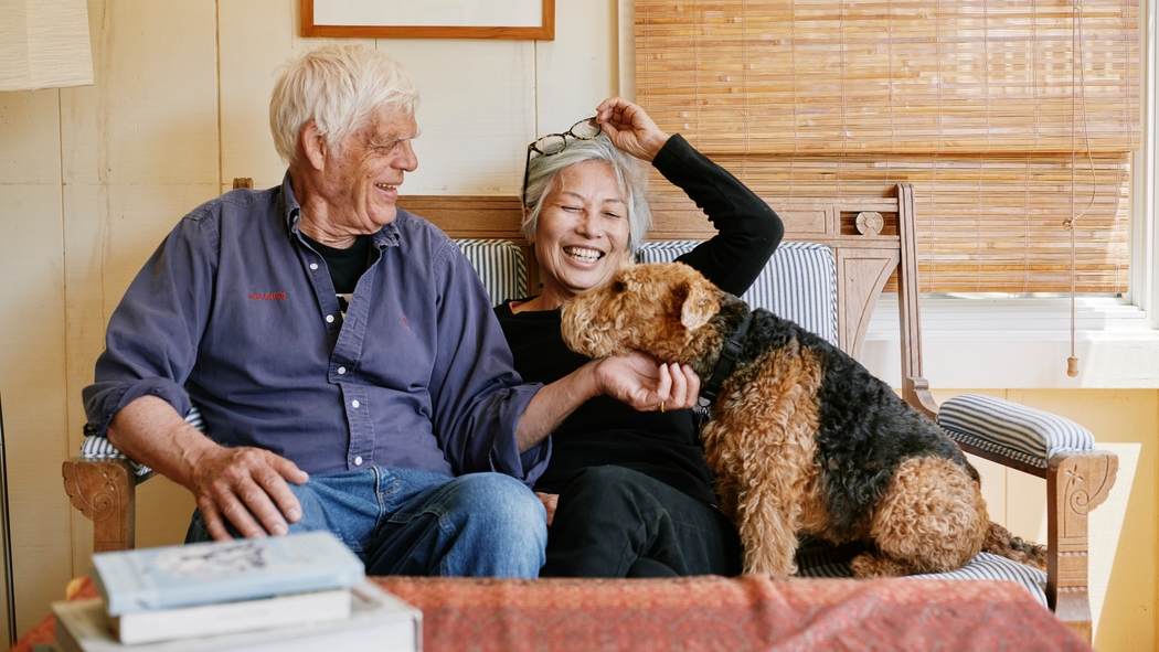A couple sits on a couch and pets a dog.