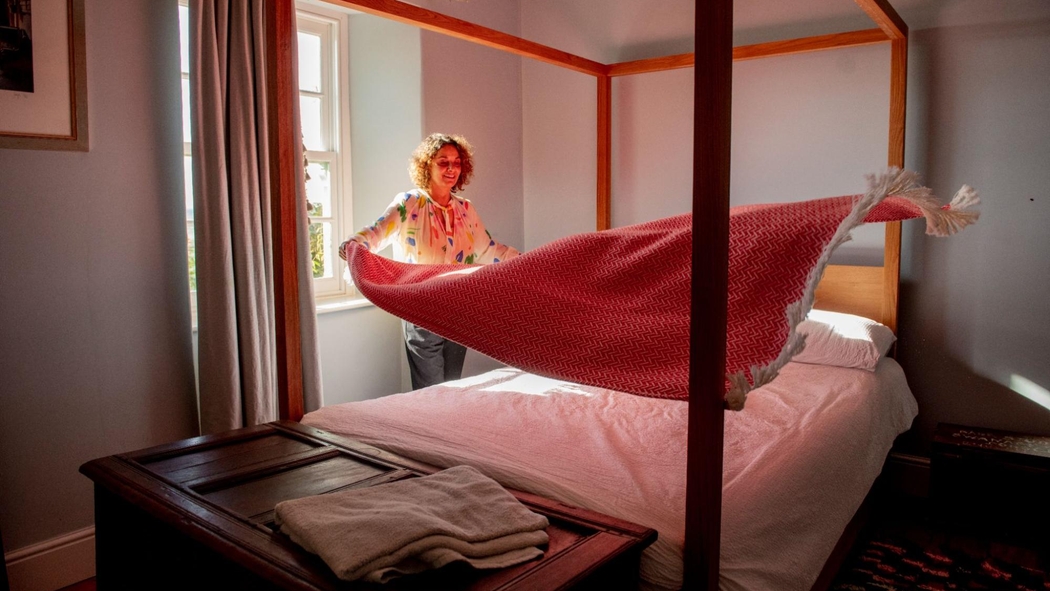 Uma anfitriã estende um cobertor com franja em uma cama com dossel. Na janela atrás dela, a cortina está aberta, deixando a luz do sol entrar no cômodo.