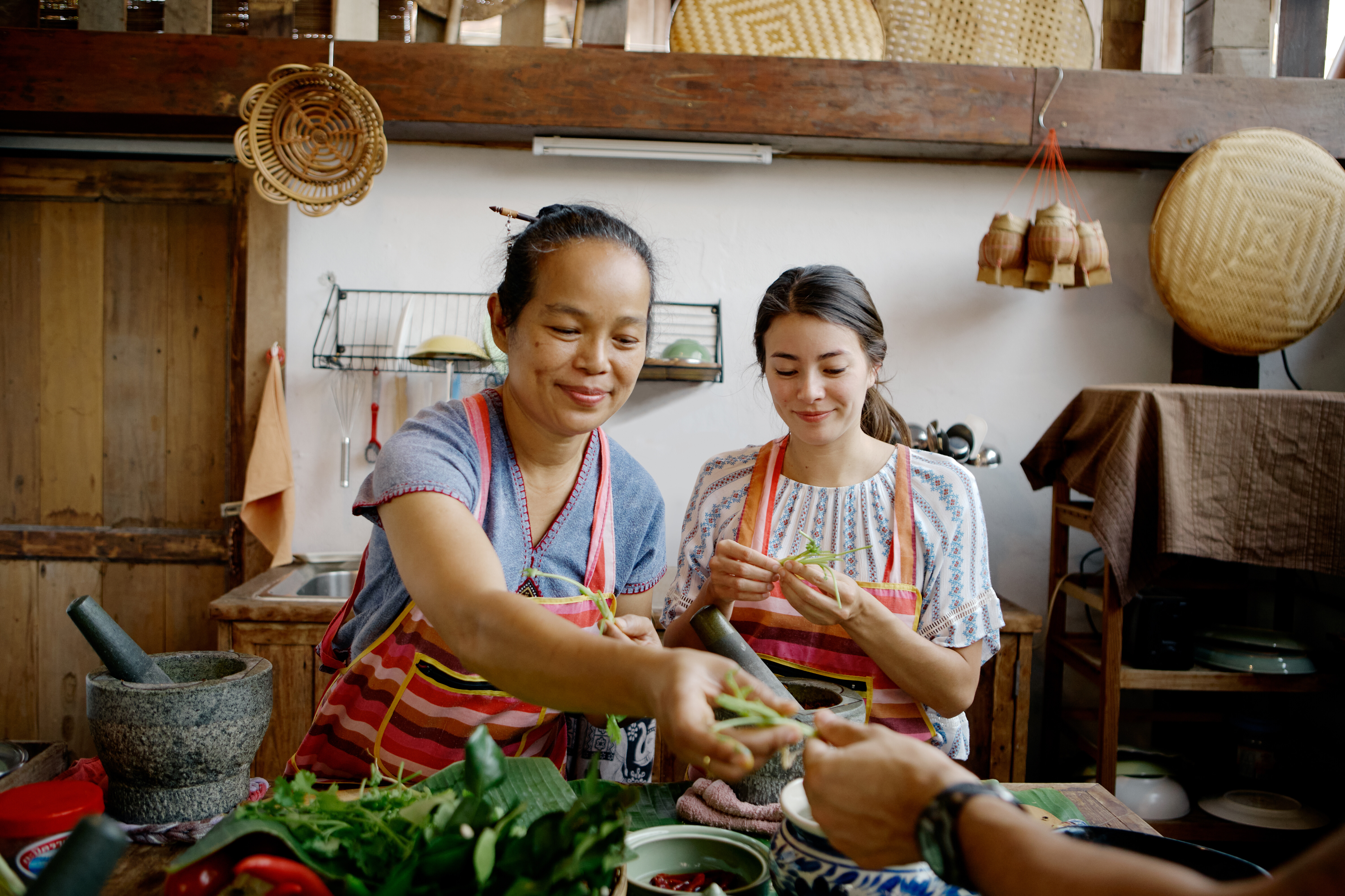 Due persone con indosso un grembiule maneggiano piante aromatiche fresche su un tavolo da cucina, dove sono presenti anche foglie di banano, peperoncini, un mortaio e un pestello.