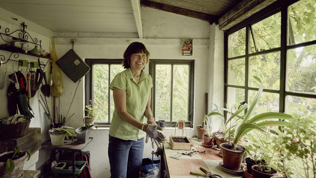 Une femme sourit en enfilant des gants de jardinage dans une pièce entourée de fenêtres et remplie de plantes en pot.