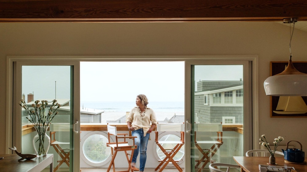 一位穿牛仔裤和奶油色衬衫的女士站在一座房屋的阳台上，这里可以俯瞰大海。