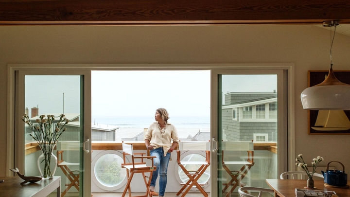 A woman wearing jeans and a cream blouse stands on the balcony of a house overlooking the ocean.