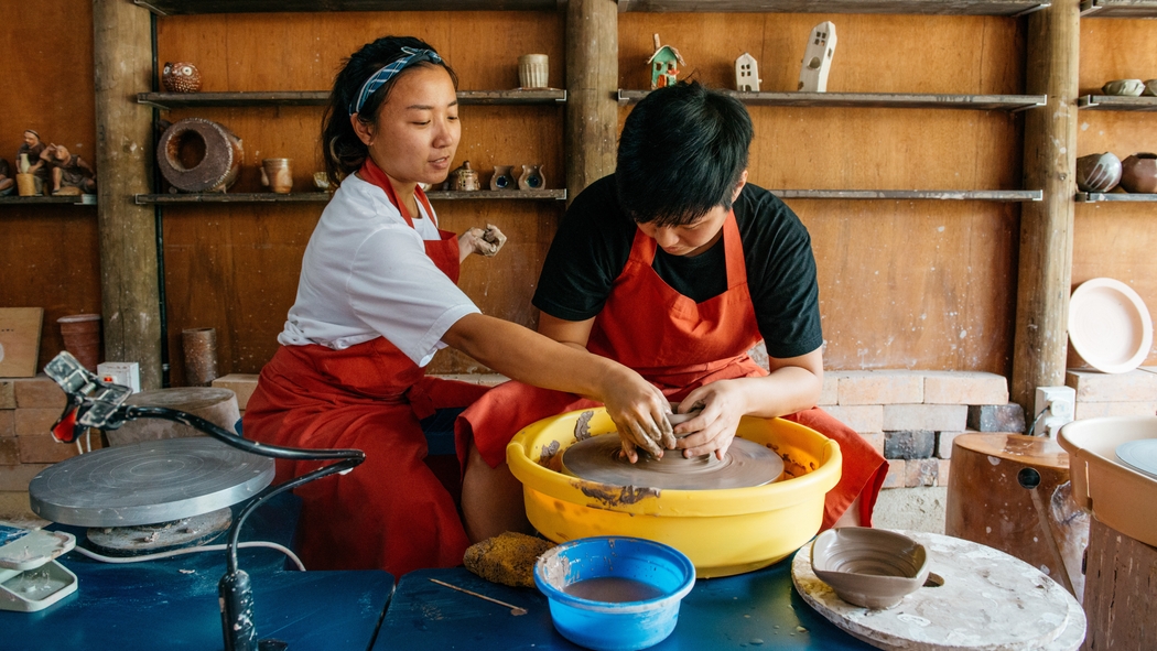 Two people wearing red aprons sit at a pottery wheel. One of them guides the other’s hands on the clay as it spins.