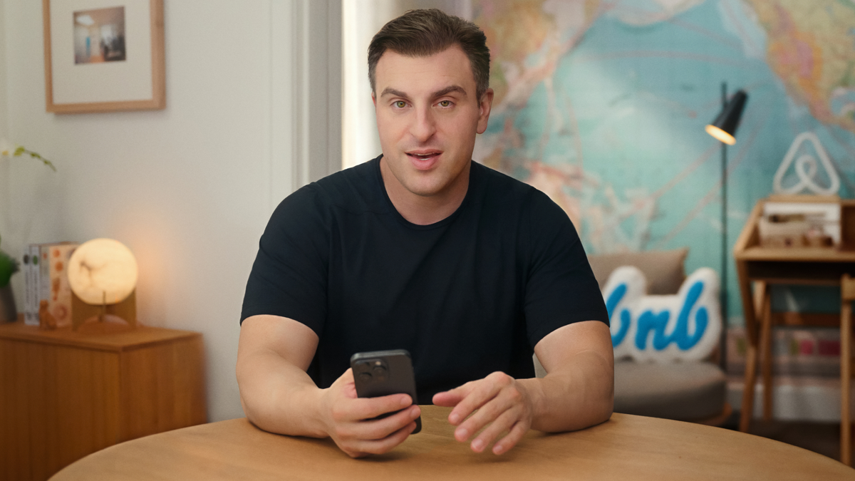 Le président d'Airbnb Brian Chesky, en t-shirt foncé, a les coudes sur une table et tient un téléphone portable dans sa main droite.