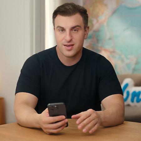 CEO Brian Chesky is wearing a dark T-shirt, resting his elbows on a table, and holding a mobile phone in his right hand.