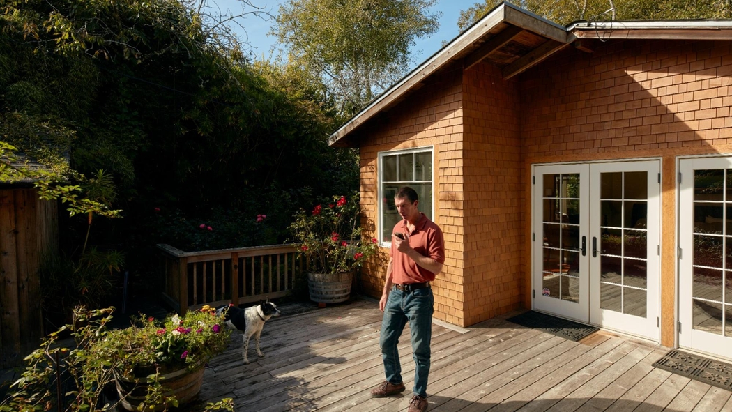 Een Airbnb-host staat op een zonovergoten terras op zijn telefoon te kijken. Naast hem staat een hond op een plekje in de schaduw.