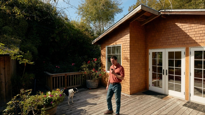 Хозяин жилья смотрит в телефон на освещенной солнцем террасе. Рядом в тени стоит собака.