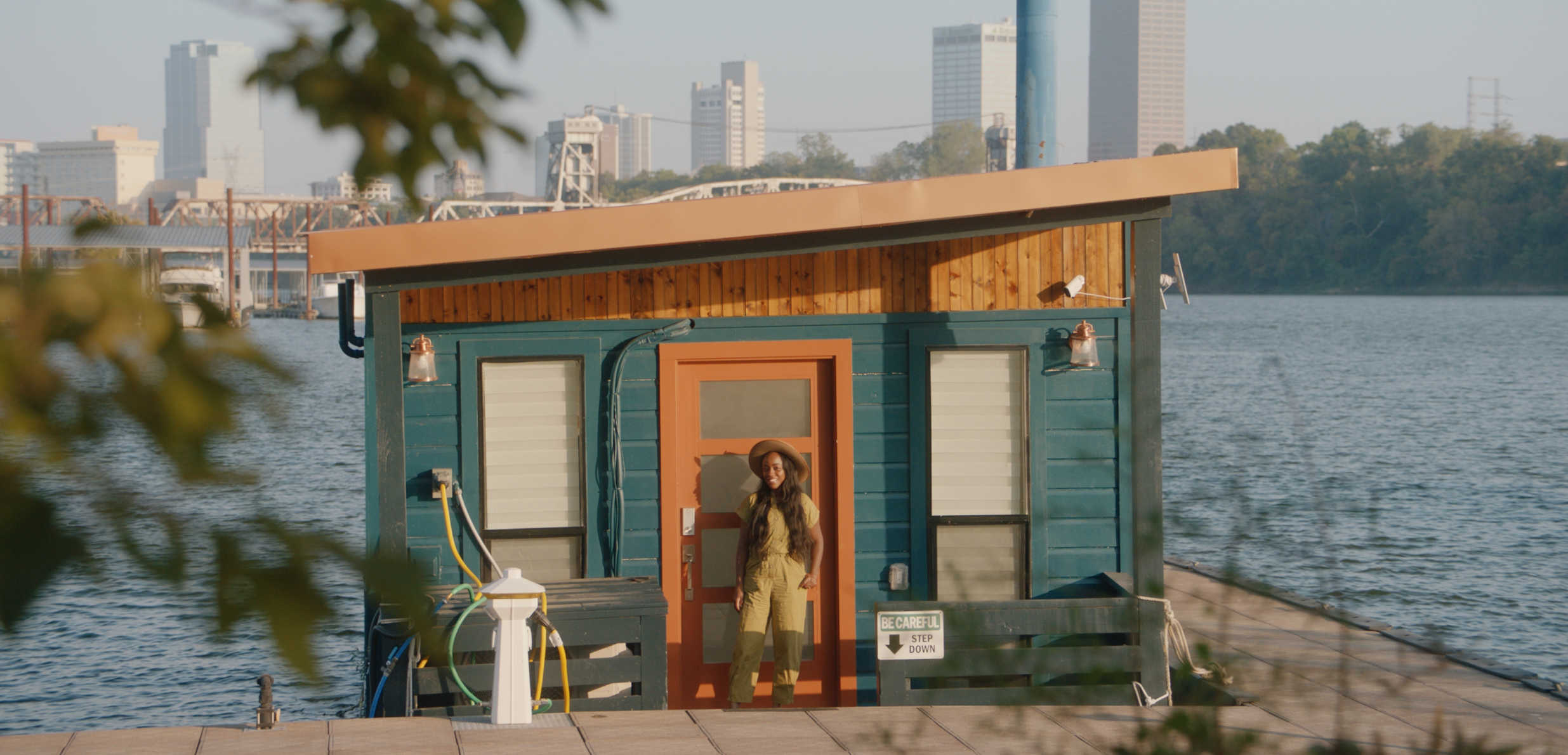 Un anfitrión sonríe frente a una casa flotante con vistas al agua, unos árboles y el paisaje urbano a lo lejos.