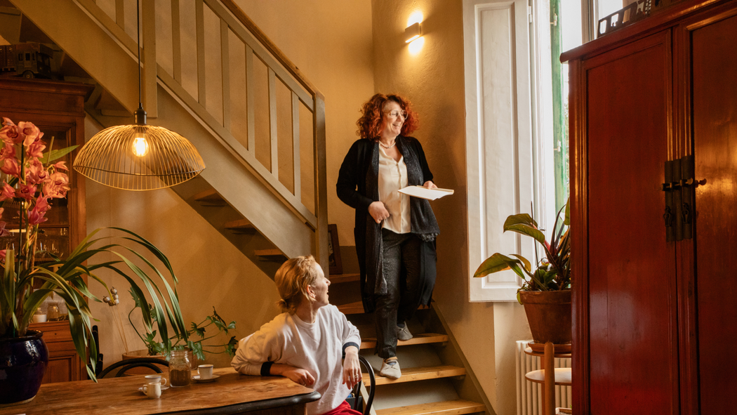 Une femme séjournant dans un logement Airbnb est assise à une table, sur laquelle est posée une tasse de café. Elle s'est retournée pour interagir avec son hôte qui descend l'escalier.