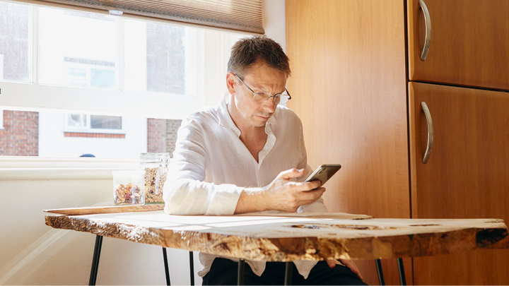 En person med glasögon sitter vid ett träbord och tittar på en smartphone. Solen skiner genom ett öppet fönster.