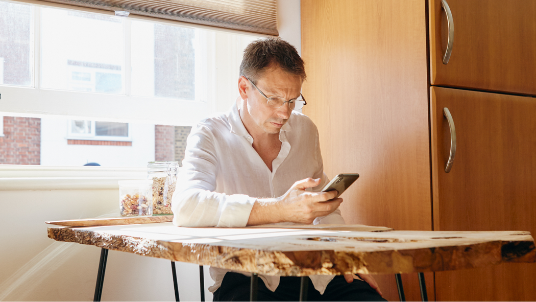 Eine Person mit Brille sitzt an einem Holztisch und schaut auf ein Smartphone. Die Sonne scheint durch ein offenes Fenster.