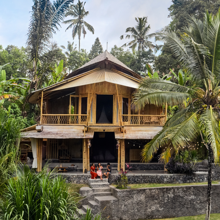 Three people sit on the front steps of a place listed on Airbnb in Bali facing large tropical trees and vegetation.