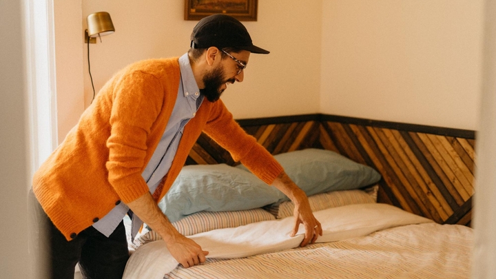 En person i orange sweater og baseballkasket smiler, mens han lægger et hvidt lagen på en dyne på en seng.