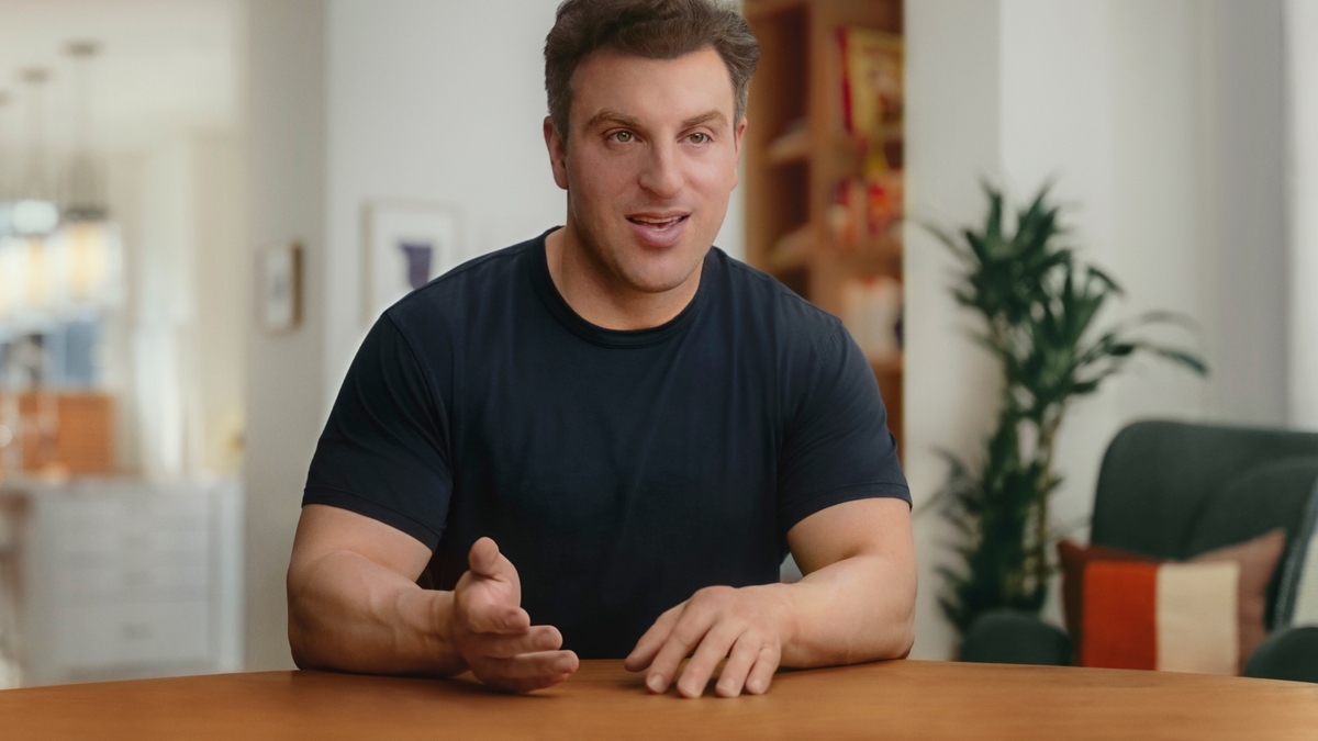 Koyu renk bir tişört giymiş Airbnb CEO'su Brian Chesky, kameraya bakarken ellerini ahşap bir masanın üzerine koyuyor.