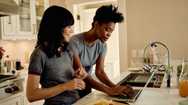 Two people look at a laptop together in a bright kitchen with a marble worktop.