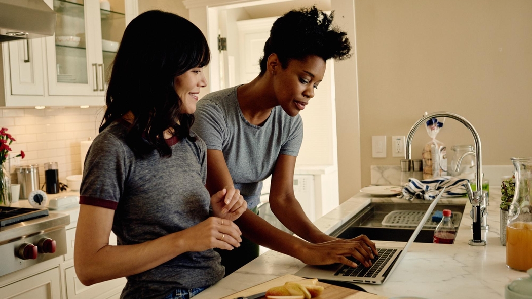 Two people look at a laptop together in a bright kitchen with a marble worktop.