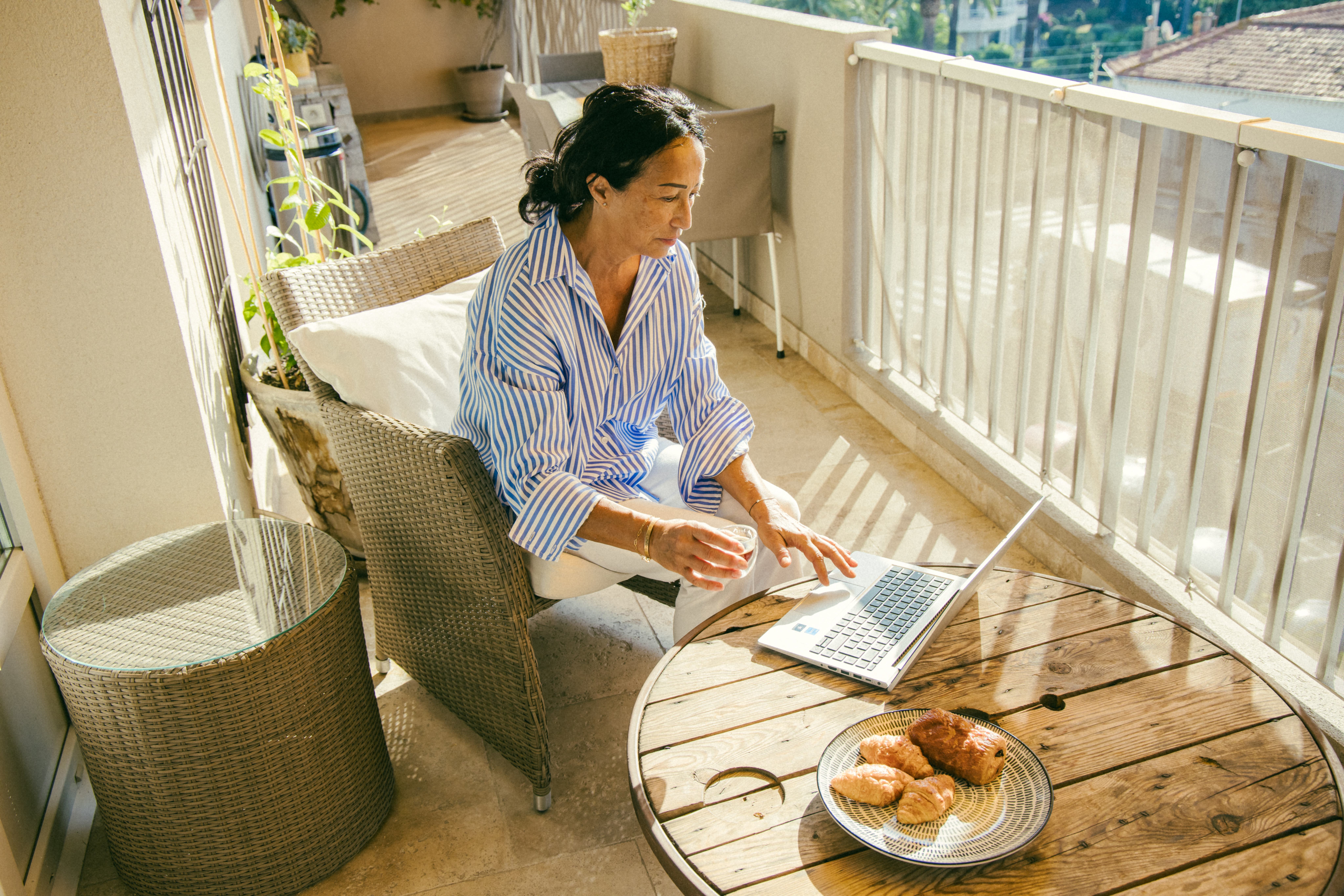 Persuna bilqiegħda ma' medja tonda tal-injam fuq veranda, qed taħdem fuq il-laptop fid-dawl tax-xemx.