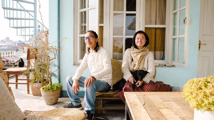 Two people sit on a balcony bench just outside large open windows near potted trees, a table, and a spiral staircase.