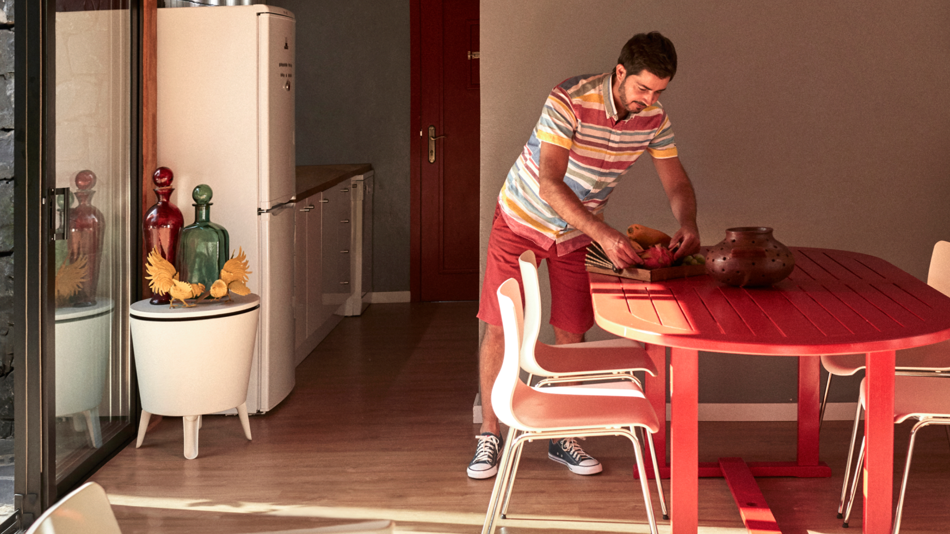 Человек в полосатой рубашке на пуговицах расставляет предметы на красном обеденном столе рядом с раздвижными стеклянными дверями.