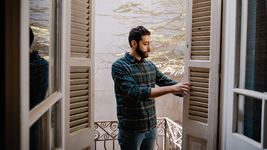 Une personne vêtue d'une chemise et d'un jean à carreaux examine les volets d'une porte-fenêtre à deux battants qui s'ouvrent sur un balcon.