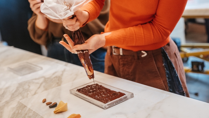 Una persona está de pie sobre una encimera de mármol blanco y exprime chocolate de un tubo en un molde rectangular transparente.