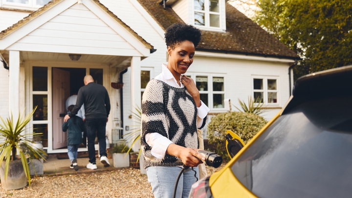 A smiling person holds an electric-vehicle charging connector next to a yellow car, outside of a white house.