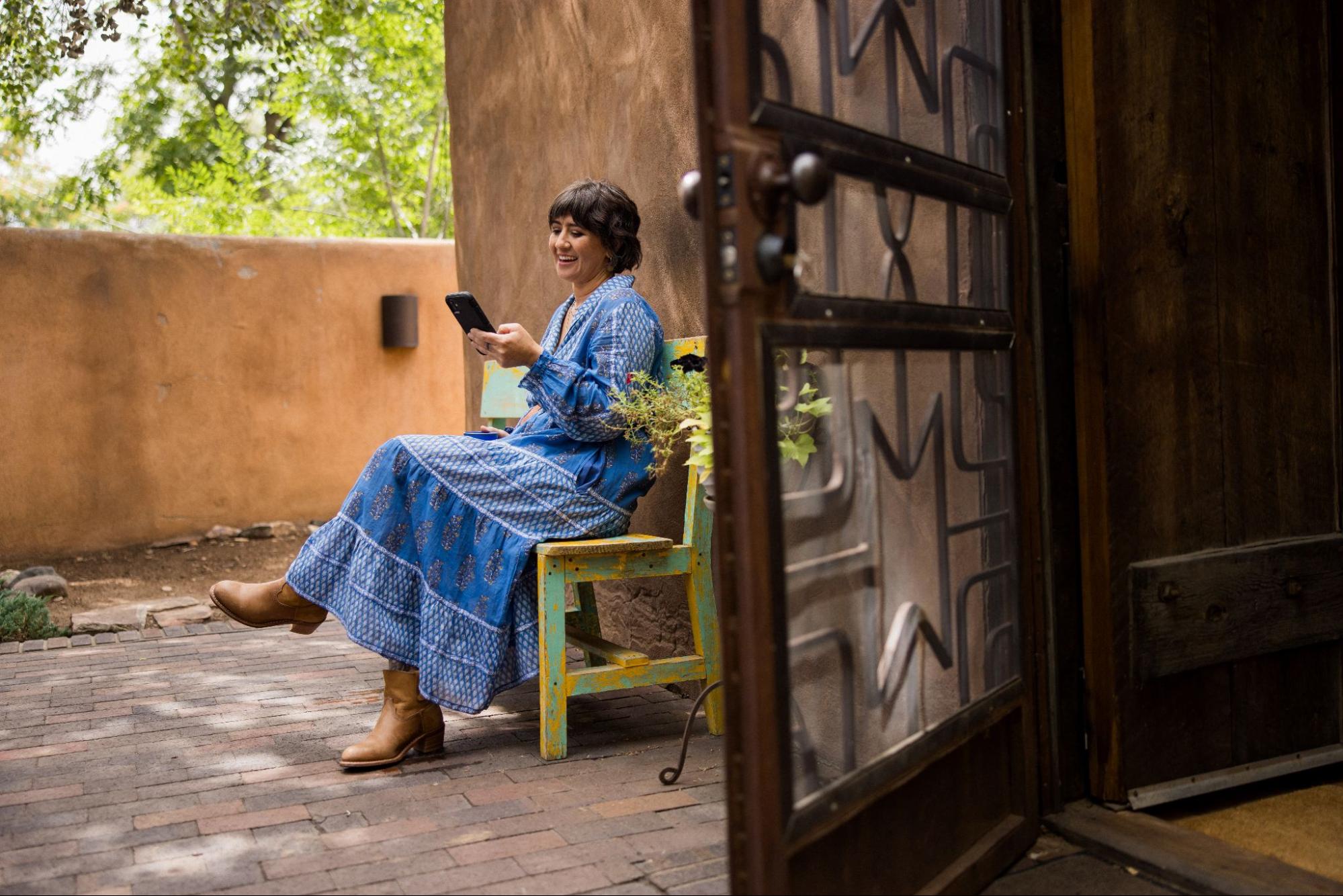 Eine Person in einem langen blauen Kleid sitzt auf einer Holzbank neben einer offenen Tür und blickt lächelnd auf ein Smartphone.