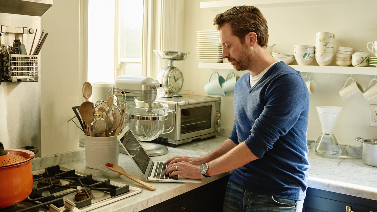 A man in a blue sweater and jeans works on a laptop on a kitchen counter.