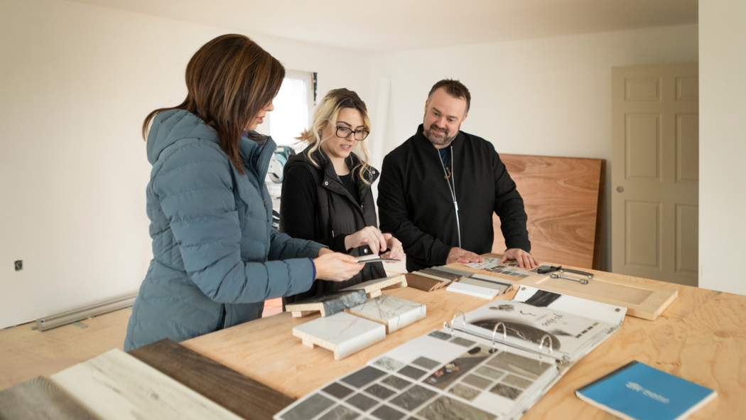 Three individuals stand at a table, looking down at sample materials for housing.