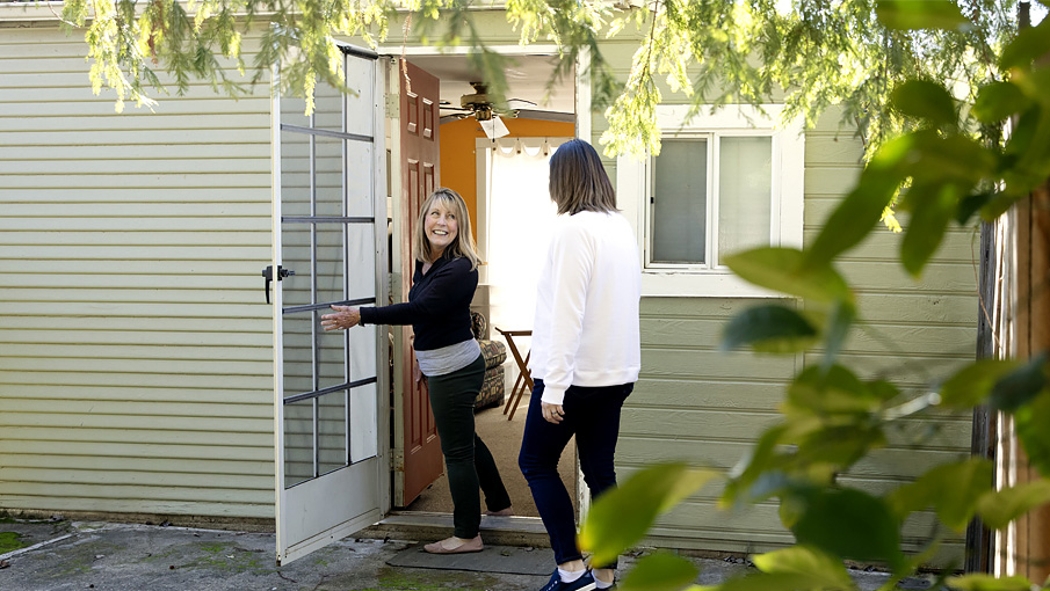 A smiling woman opens the front door, welcoming another woman inside.
