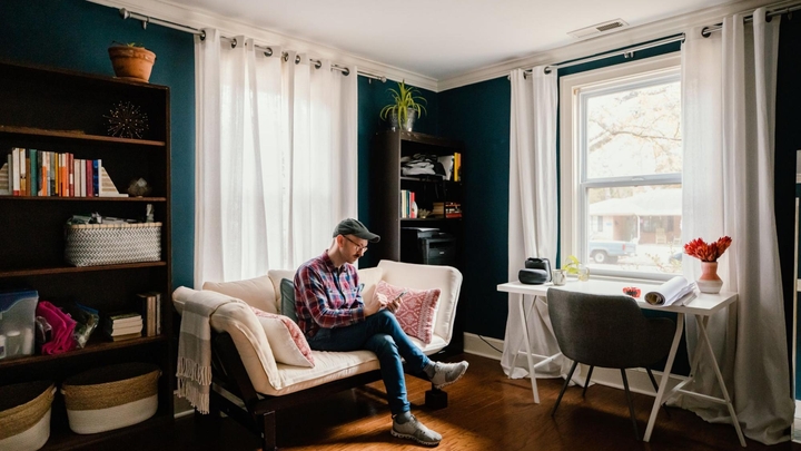 Un adulto sentado con las piernas cruzadas en medio del sillón de una sala mirando un teléfono inteligente en sus manos.