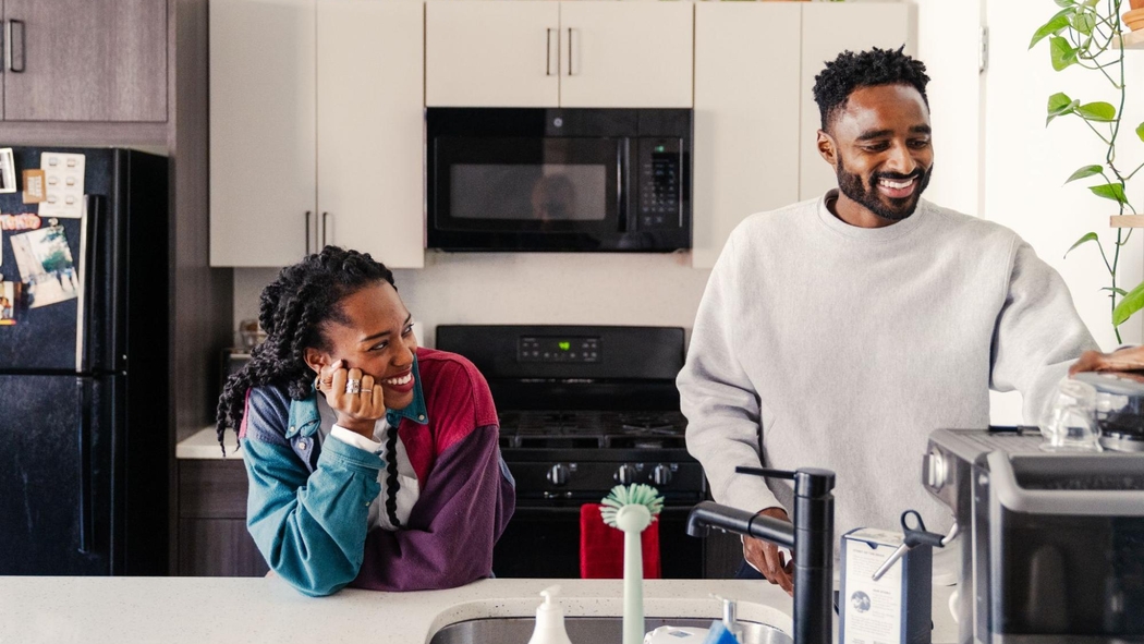 兩個人面帶微笑，站在一間風格現代且設備齊全的廚房中，其中一人正在準備煮咖啡。