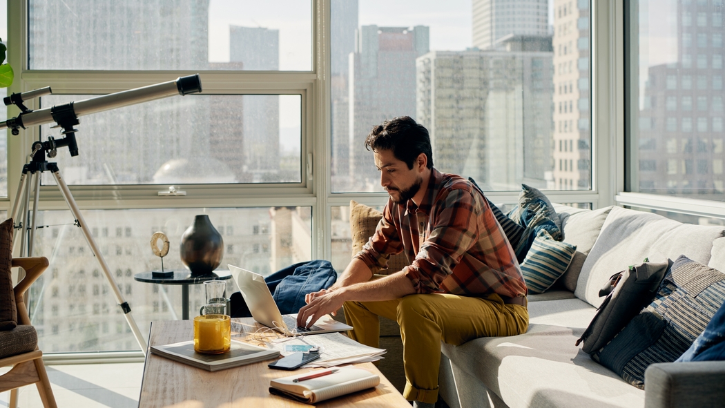 Человек печатает на ноутбуке, сидя на диване в квартире с панорамным остеклением. Открывается вид на город и высотные здания.