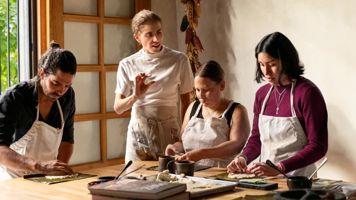 Four people wearing aprons are standing around a kitchen island, spreading masa on banana leaves. A fifth person guides them.