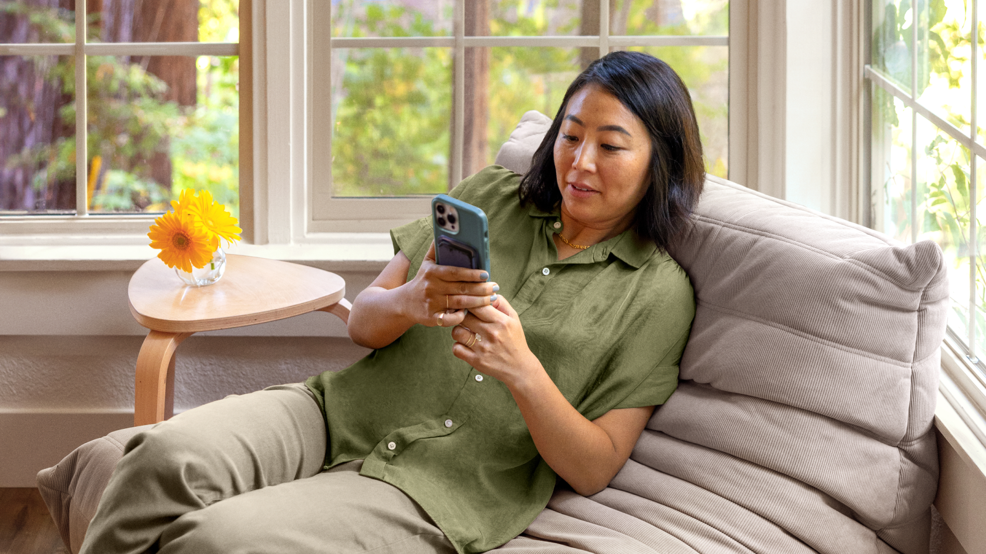 Ένα άτομο με ένα λαδί πουκάμισο με κουμπιά κοιτάζει ένα smartphone και ξαπλώνει σε μια μπεζ καρέκλα δίπλα σε παράθυρα με θέα στα δέντρα.