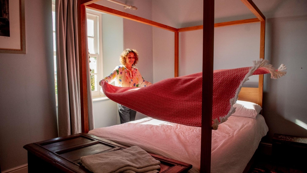 A person makes a four-poster bed, shaking out a red blanket while sunlight streams in through the window. 