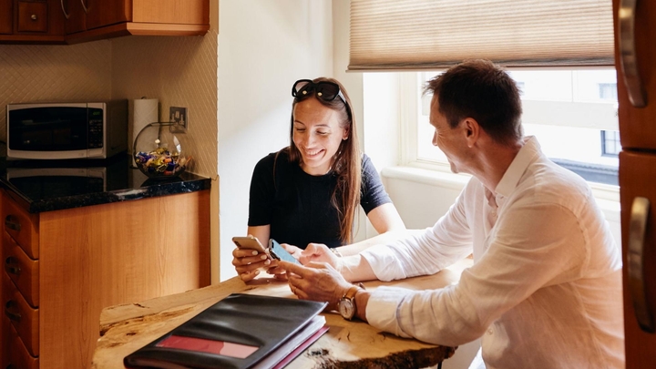 Dos personas sonríen y escriben en sus celulares mientras están sentadas en una mesa de cocina pequeña hecha de madera.