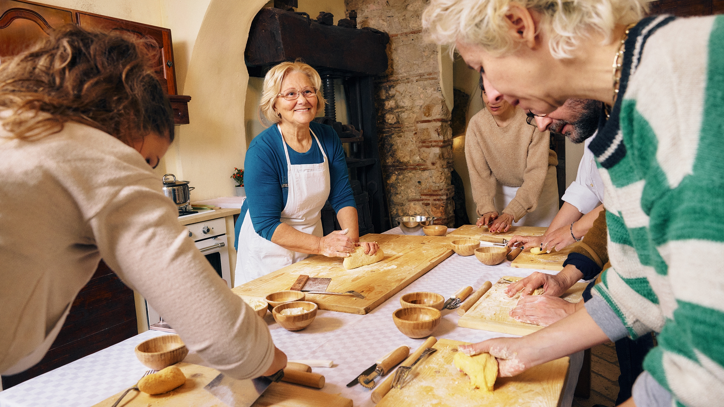 คน 5 คนยืนอยู่รอบโต๊ะเตรียมอาหารและนวดแป้งบนเขียงไม้ของแต่ละคนที่โรยด้วยแป้ง