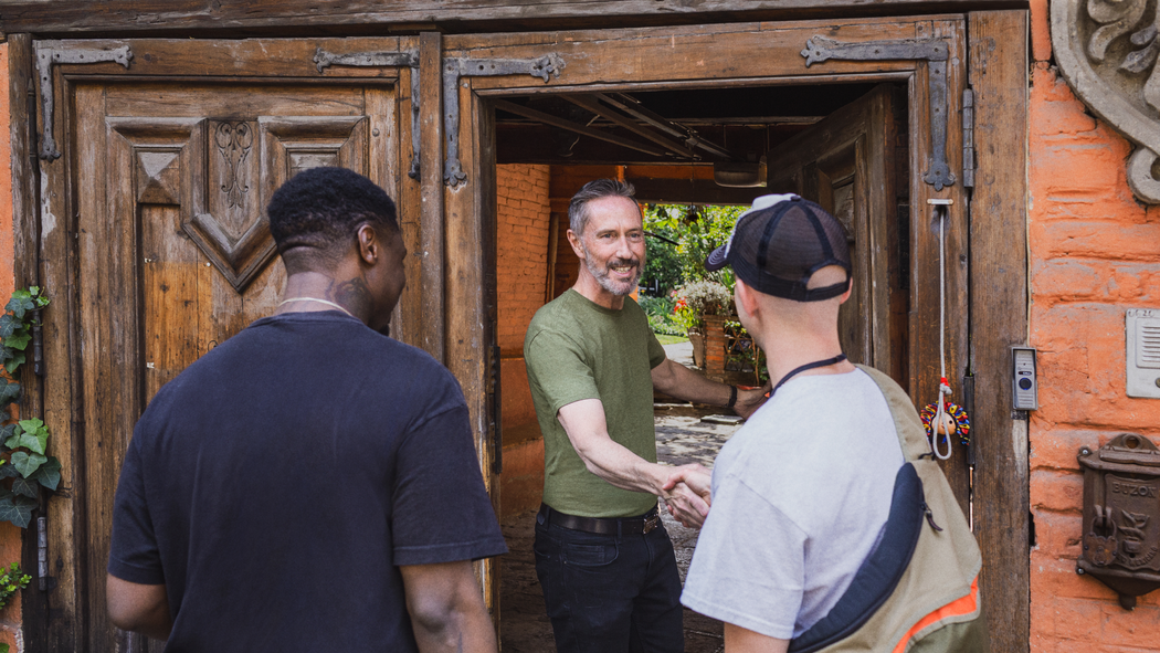 Superhost Werner shakes hands with one of two guests arriving at his home. The open door reveals brick walls and a garden.