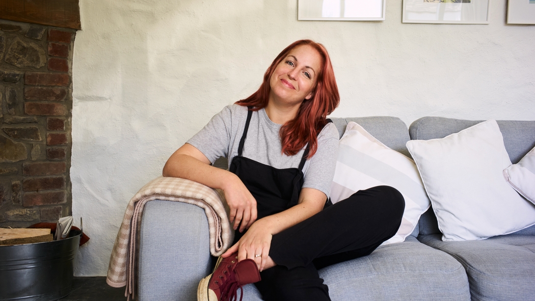 En kvinna med rosa hår sitter på en grå soffa i en stuga med gräddvita väggar och en öppen spis i tegel.