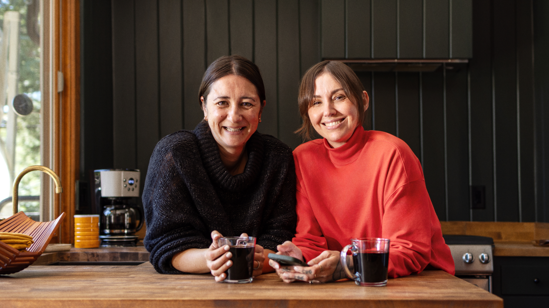 Two people smile and lean on a kitchen counter with mugs of coffee in front of them.