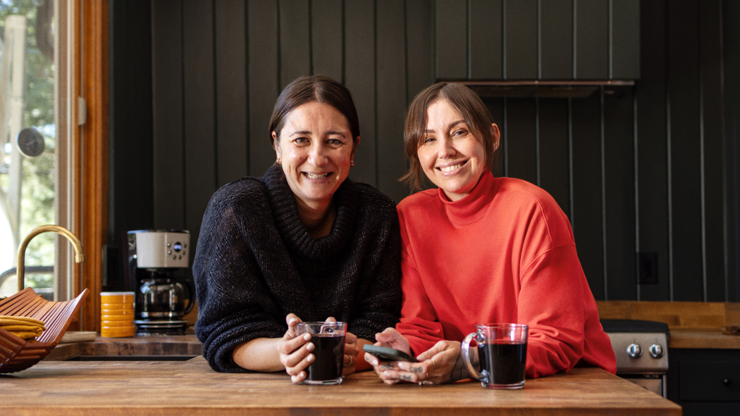 Two people smile and lean on a kitchen counter with mugs of coffee in front of them.