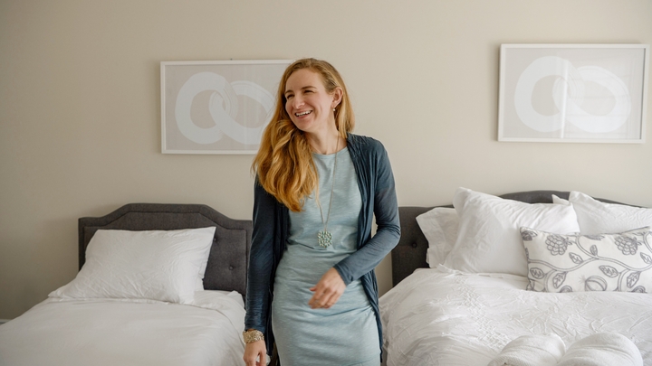 A blond woman in a blue dress stands smiling in a bedroom between two beds with white linens.
