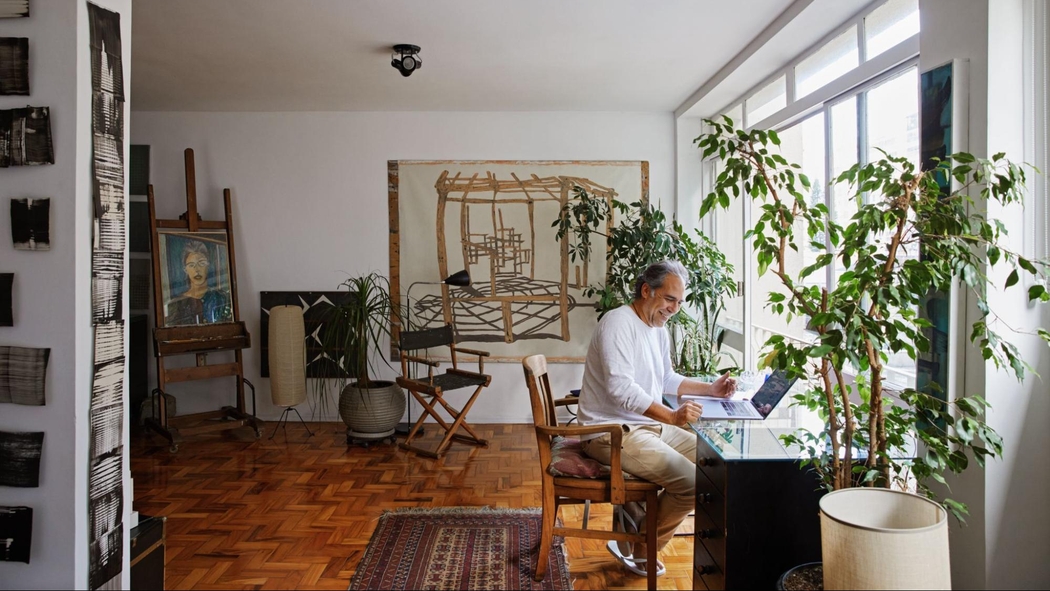 Een glimlachend persoon met grijs haar zit aan een bureau met een laptop in een kamer met planten, grote ramen en kunst.