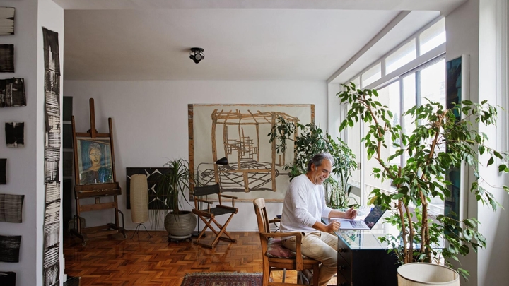 Umntu oncumileyo oneenwele ezingwevu uhleli kwidesika ne-laptop kwigumbi elinezityalo, iifestile ezinkulu nemizobo.