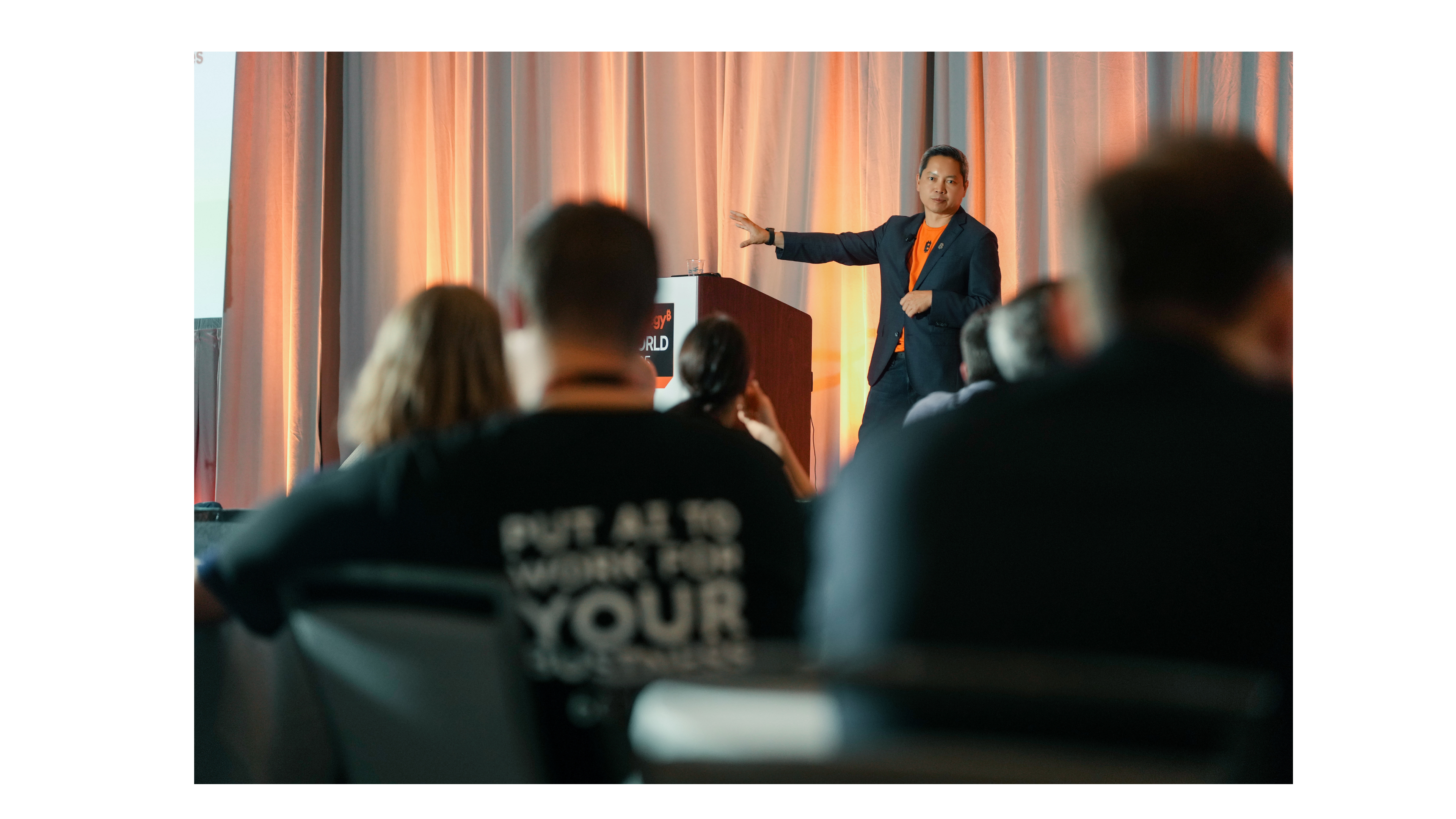Strategy Ai Transformation Forum speaker and CEO, Phong Le, in orange branded attire gestures to an engaged audience during a live enterprise AI presentation, with warm stage lighting and a wooden podium displaying Strategy World Tour branding.