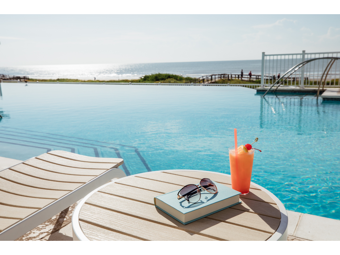 SSR-Poolside-Drinks	Poolside table with book, sunglasses, and tropical drink by ocean.